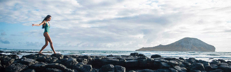 Person walking along rocks at the beach with a mountain in the background