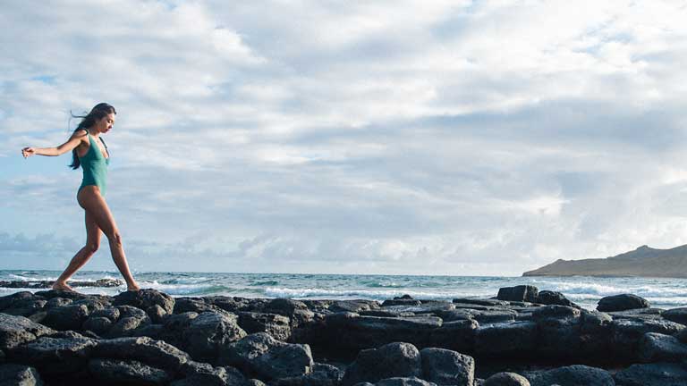 Person walking along rocks at the beach with a mountain in the background